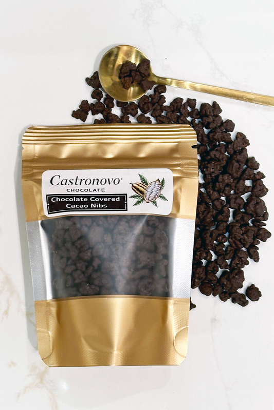 A gold-sealed package of Castronovo brand dark chocolate covered cacao nibs, with a spoonful of the product resting on top of the scattered nibs.