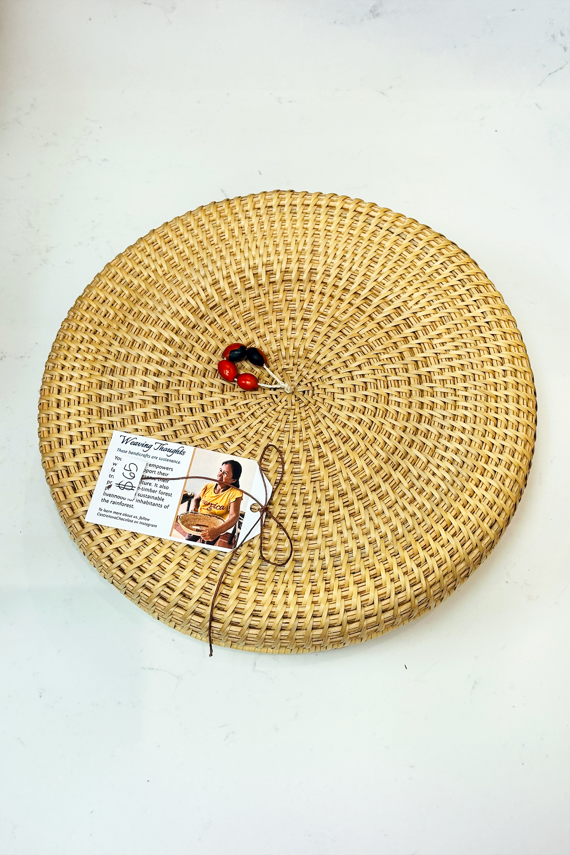 A round, woven basket made of natural materials by the women of the Piaroa indigenous group, with a tag attached displaying a picture and information about the product.