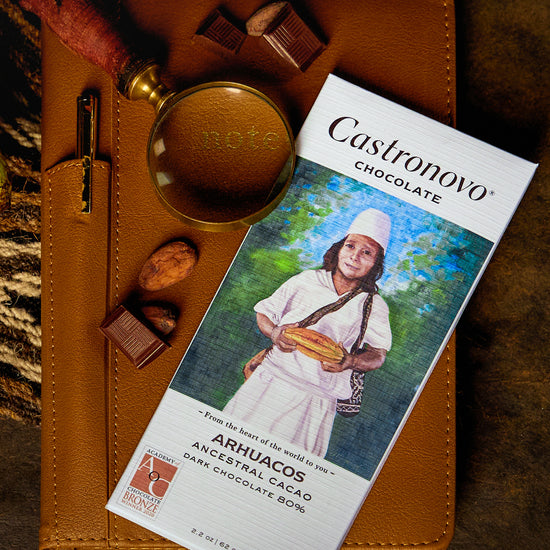 A bar of Arhuacos 80% dark chocolate by Castronovo Chocolate, placed on a surface with cocoa beans and a magnifying glass.