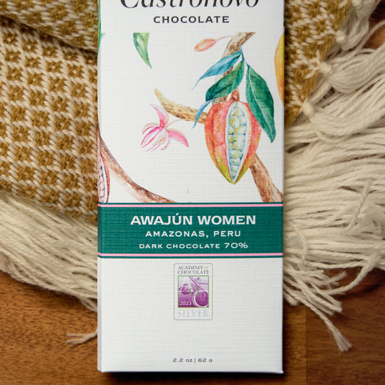 A chocolate bar with the label 'CASTRONOVO CHOCOLATE' and 'Awajun Women Peru 70% Dark'. The packaging is predominantly white and green with an illustration of a cacao tree and flowers.