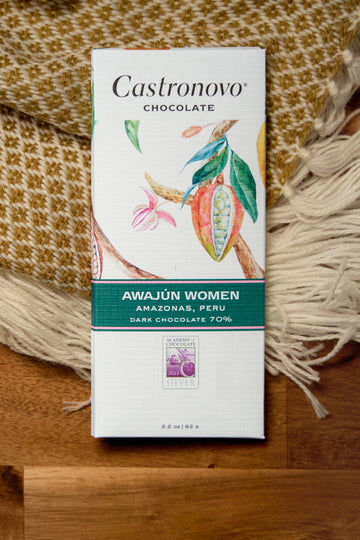 A chocolate bar with the label 'CASTRONOVO CHOCOLATE' and 'Awajun Women Peru 70% Dark'. The packaging is predominantly white and green with an illustration of a cacao tree and flowers.