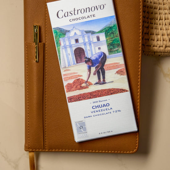 A bar of Castronovo Venezuela 72% Dark chocolate placed on top of a brown notebook with an illustration of a person on the cover, set on a kitchen counter.