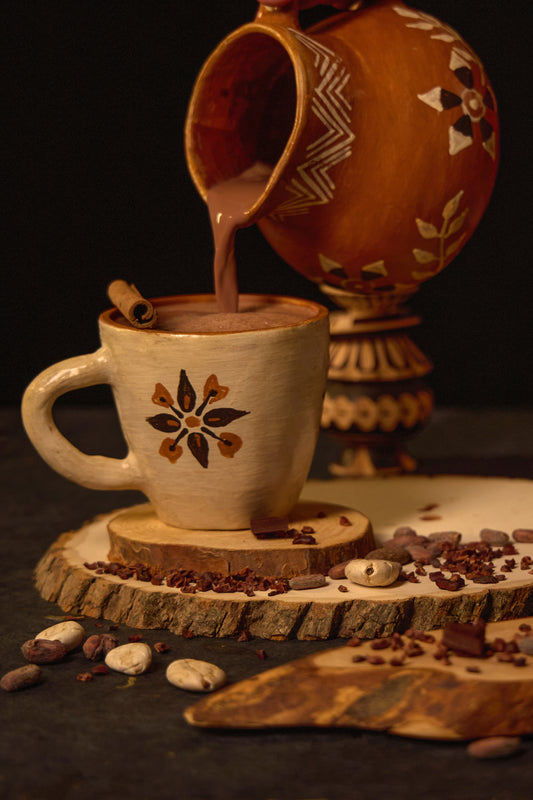 A mug of sipping chocolate with a patterned design, accompanied by scattered cocoa beans and a cinnamon stick, with a traditional ceramic pot in the background.