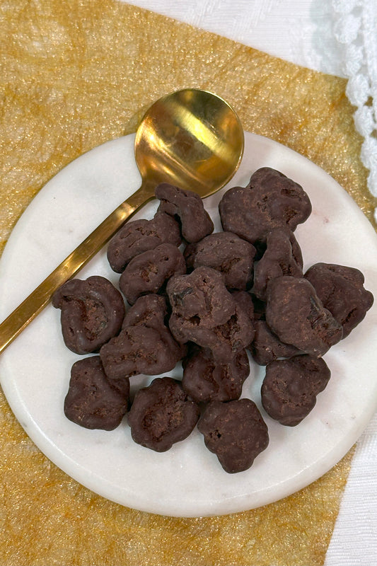 A plate of dark chocolate covered Montmorency cherries with a golden spoon on the side, placed on a yellow napkin.