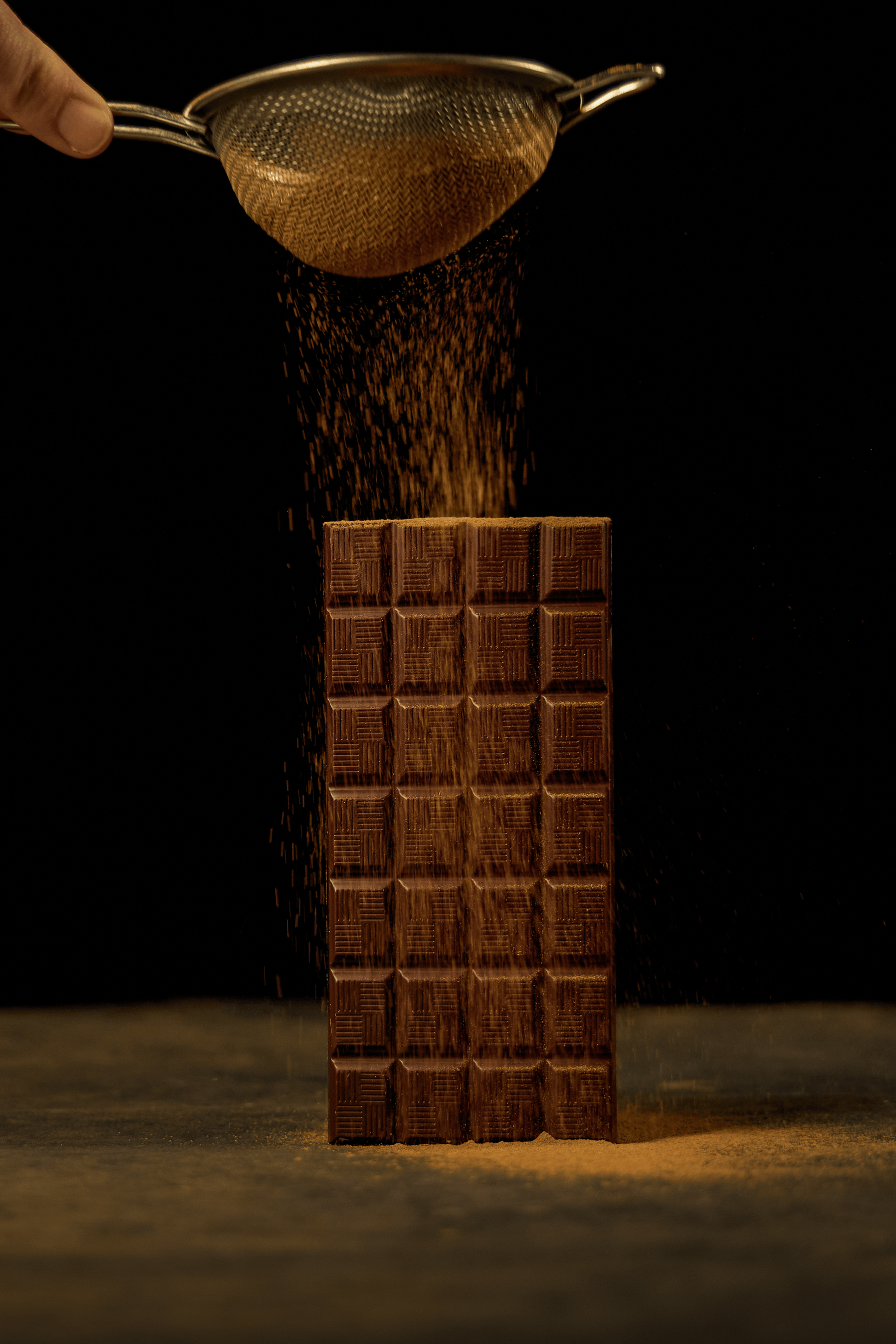 Photograph of chocolate powder being sifted onto upright chocolate bar against dark background
