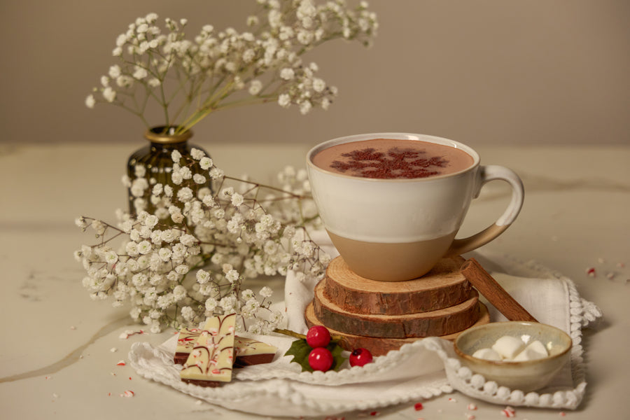 Image of hot sipping chocolate in mug on tan background surrounded by flowers, peppermint chocolate bark, and marshmallows