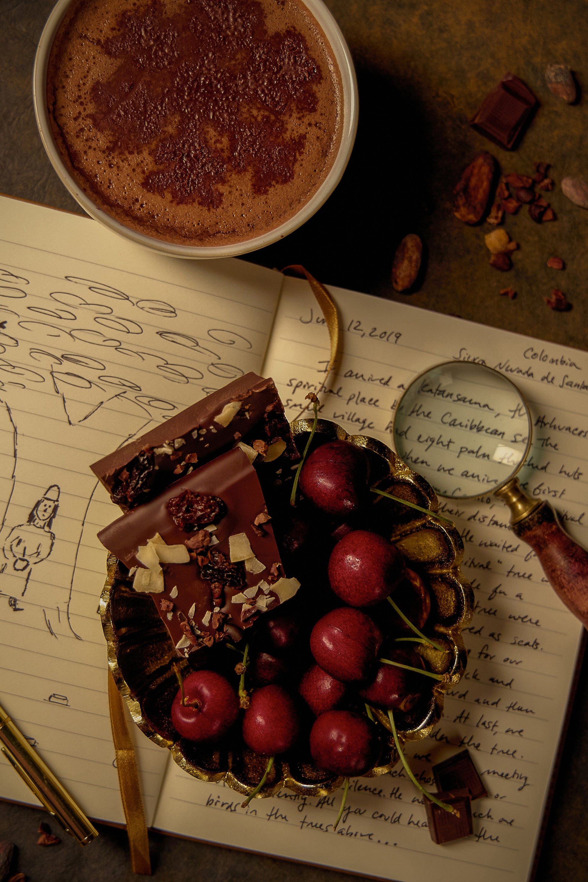 A picture of cherry, coconut, cacao nib bark in a bowl of cherries on top of a journal record about the Sierra Nevada de Santa Marta, Colombia. Beside the bowl is a cup of hot drinking chocolate.