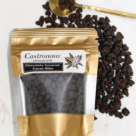 A gold-sealed package of Castronovo brand dark chocolate covered cacao nibs, with a spoonful of the product resting on top of the scattered nibs.