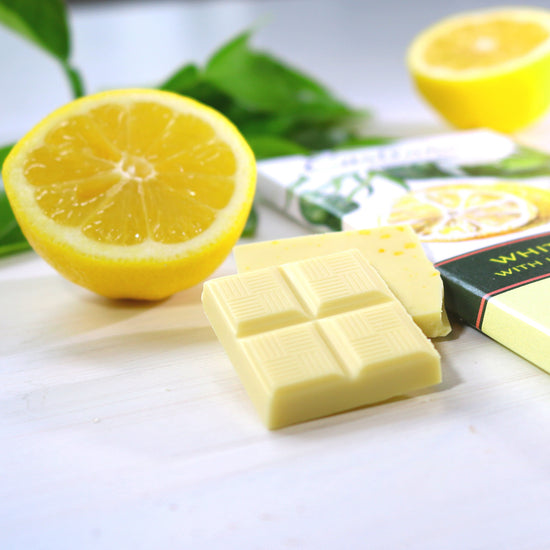 Zoomed in photo of piece of Castronovo brand white chocolate with lemon and lemon sea salt on a white cloth surface, accompanied with sliced lemon 