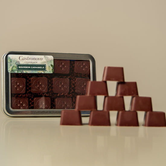 A tin of chocolate bonbons placed on a table with several bonbons arranged outside the tin, coated in dark chocolate and caramel.