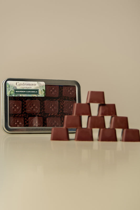 A tin of chocolate bonbons placed on a table with several bonbons arranged outside the tin, coated in dark chocolate and caramel.