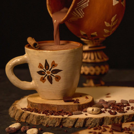 A mug of sipping chocolate with a patterned design, accompanied by scattered cocoa beans and a cinnamon stick, with a traditional ceramic pot in the background.