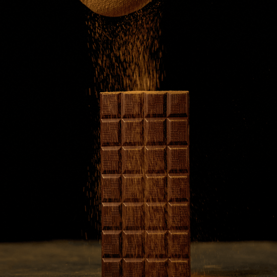 Photograph of chocolate powder being sifted onto upright chocolate bar against dark background 