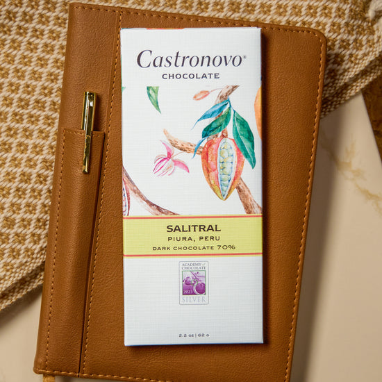 A piece of dark chocolate with a label that reads 'Castronovo Chocolate', 'SALITAL', and '70% BANCHO COLTS'. The chocolate is placed on a textured surface, possibly a fabric, with a pen and a small floral decoration beside it.