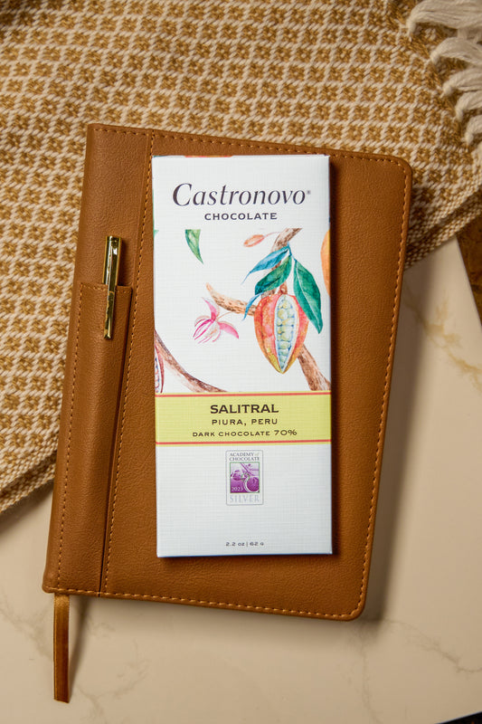 A piece of dark chocolate with a label that reads 'Castronovo Chocolate', 'SALITAL', and '70% BANCHO COLTS'. The chocolate is placed on a textured surface, possibly a fabric, with a pen and a small floral decoration beside it.
